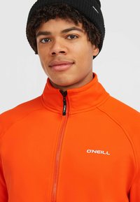 Oranje ritsjack met een hoge kraag, voorzien van een zwarte rits en een wit logo op de borst. De stof lijkt glad met een aangesloten ontwerp.