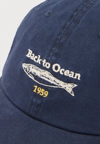 Μπλε σκουφί με λευκό κεντημένο ψάρι και κείμενο "Back to Ocean" πάνω από το ψάρι, και κίτρινο "1959" κάτω από το ψάρι.