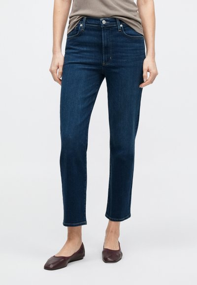 Sötétkék farmernadrág vékony szabással, középmagas derékkal, öt zsebbel és finom varrással. Bordó balerinacipővel viselve.