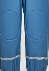 Mėlynos vandeniui atsparios kelnės su paminkštintomis kelio vietomis, pasižyminčios lygiu paviršiumi ir atspindinčiomis sidabrinėmis juostelėmis ant pundų.