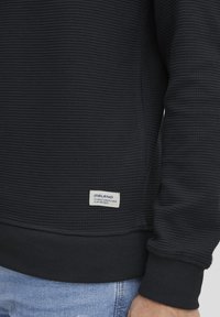 Schwarzer, strukturierter Sweatshirt mit Rippmuster, ausgestattet mit einem kleinen Etikett unten links und einem taillierten Bündchen.