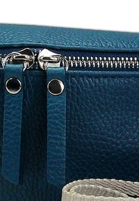 Blauwe leren clutch met textuur, zilveren ritssluiting, gedetailleerde trekkers en contrasterende lichte stofaccenten. Close-up van de stiksels en hardware.