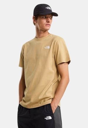 Joven con camiseta beige de The North Face, gorra negra y pantalones cortos negros, con las manos en los bolsillos, mirando hacia un lado.