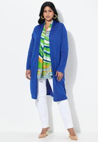 Koninklijk blauwe lange cardigan in een textuurgebreide stof, met een open voorkant, gecombineerd met een meerkleurige gestreepte tuniek en witte broek. Beige hakken maken de look af.