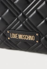 Črna prešita usnjena torba z zlatim logotipom. Ima diagonalne šive in kovinske vložke ter prikazuje oblazinjen tekstur.