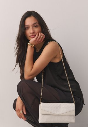 Young woman with long dark hair squatting, wearing a black sleeveless top and sheer black pants, holding a white textured purse with gold chain strap.
