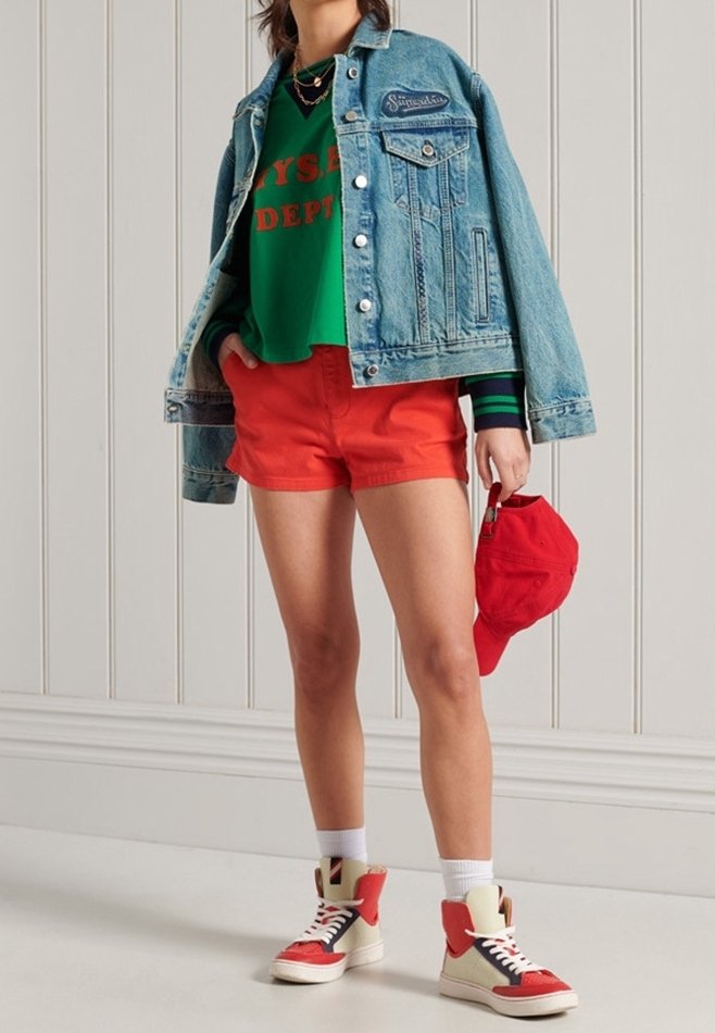 Denim jas met zakken, groene sweatshirt met rode tekst, rode shorts, wit-en-rood hoge sneakers, en een rode pet.