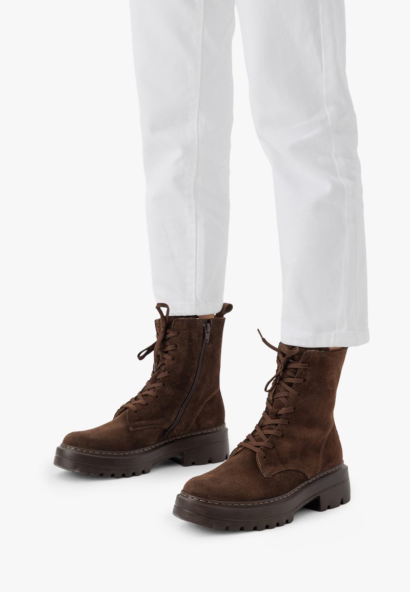 Stivaletti in suede marrone con suola spessa, lacci anteriormente e cerniera laterale. Abbinati a pantaloni bianchi. Finitura testurizzata e design resistente.