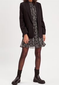 Veste noire portée sur une robe noire et blanche à motifs avec des manches à volants, associée à des bottines noires et des collants noirs transparents.