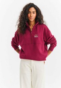 Bluza bordowa na zamek z wysokim kołnierzem i haftowanym logo "Naoko Studio". Miękki materiał, luźny krój i ściągacze na rękawach.