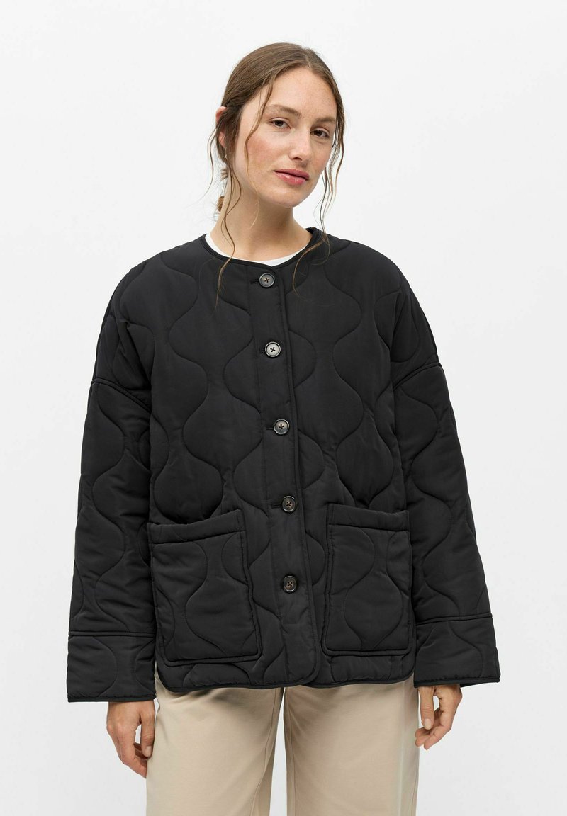 Femme portant une veste matelassée noire à boutons avec deux poches avant et un pantalon beige, debout devant un fond blanc uni.