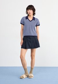 Polo shirt à motifs bleu marine et blanc avec des manches courtes, associé à une jupe évasée noire. Des sandales à talon bas beige et des chaussettes rayées sont portées.