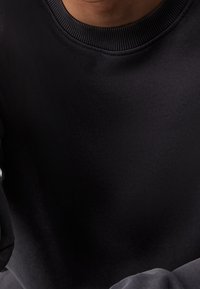 Hombre con un suéter negro de cuello redondo, visible desde el cuello hasta la parte superior de la cintura, sentado con los brazos descansando frente a él.