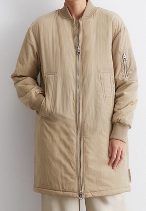 Beige long bomber jacket with ribbed cuffs, front pockets, zipper pocket on sleeve, and double zipper closure worn by a person.
