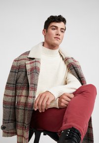 Abrigo de cuadros de lana en gris, beige y rojo, con un suéter de cuello alto color crudo y pantalones burdeos. Botas negras con cordones y acentos texturizados.