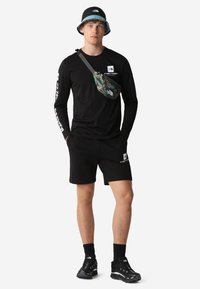 Maglietta nera a maniche lunghe e pantaloni corti con il logo The North Face, abbinati a una borsa a tracolla camouflage e sneakers nere. Cappello a secchiello con bordo azzurro chiaro.