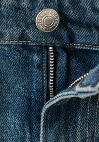 Detalle del zipper y botón de unos vaqueros de mezclilla; tela azul, costuras en contraste, herrajes de metal plateado y un zipper suave con dientes visibles.