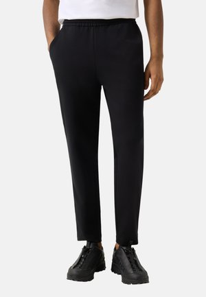 Mand iført sorte slim-fit bukser med elastisk taljebånd og sorte sneakers, stående med hænderne afslappet ned langs siderne.