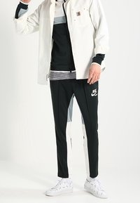 Sportoutfit med en svart zip-up jacka över en randig skjorta, kombinerad med svarta och grå byxor, samt vita sneakers. Innehåller en logotyp på byxorna.