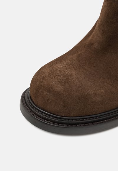 Stivaletto in suede marrone con punta rotonda e suola in cuoio nera cucita; texture morbida e design senza soluzione di continuità senza hardware visibile.