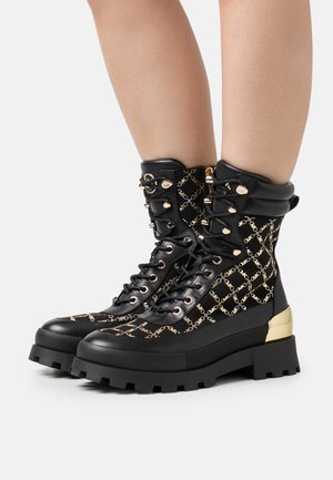 Sorte snørestøvler med ankelhøjde, der har et mønster af guldkæder, guldfarvede metaldetaljer og en kraftig sort sål med en metallic guldfarvet hældetalje.