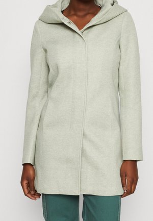 Woman wearing light gray hooded coat with concealed front closure and green pants, shown from neck to hips.