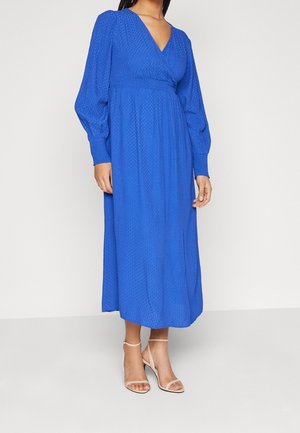 Femme portant une longue robe bleue texturée avec des manches longues bouffantes et des sandales à talons hauts beiges à lanières, debout devant un fond uni.