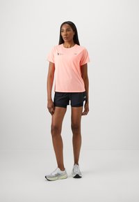 T-shirt de sport à manches courtes, rose clair avec logo, associé à un short noir et des chaussures de course grises avec des accents blancs et jaunes.