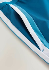 Gros plan sur un vêtement turquoise avec une bande blanche le long du bord, montrant la texture du tissu et les détails de la couture.