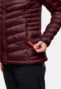 Giacca isolante bordeaux con baffi orizzontali. Presenta una tasca con zip dotata di un tirante arancione per il materiale e un accento verde.
