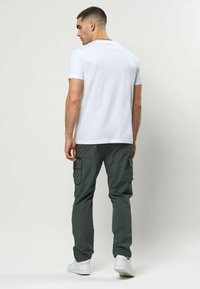 Homme portant un t-shirt blanc uni, un pantalon cargo vert foncé et des baskets blanches, debout de dos sur un fond blanc.