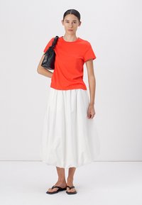 Tricou din bumbac roșu aprins, asortat cu o fustă albă, vaporoasă. O geantă de mână din piele neagră și papuci negri completează ținuta.