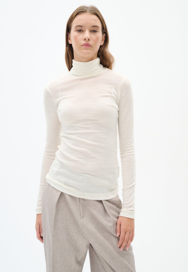 Long-sleeve turtleneck shirt in light beige, soft, sheer fabric. Features a fitted silhouette and a smooth texture, paired with high-waisted trousers.