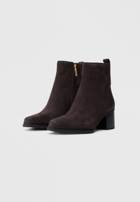 Stivali alla caviglia in suede marrone con punta affusolata, tacco a blocco medio e dettaglio con zip dorata sul lato. Texture liscia, design elegante.