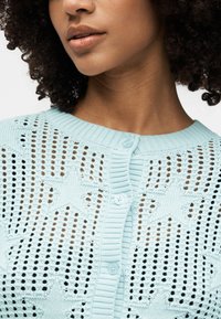 Κοντινό πλάνο ενός ατόμου που φοράει ένα ανοιχτό μπλε πλεκτό ζακέτα με σχέδια αστεριών και κουμπιά κατά μήκος του κέντρου.