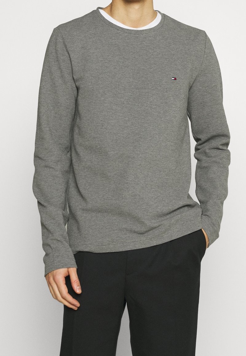 Grauer Pullover mit langen Ärmeln, geripptem Textur und Rundhalsausschnitt, mit einem kleinen Logo auf der Brust, kombiniert mit dunklen Hosen.