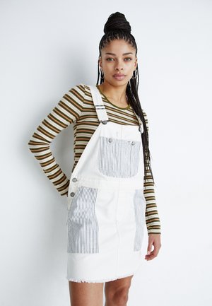 Vestito in denim bianco con tasche a righe, orlo grezzo e minuteria in argento. Abbinato a una camicia a maniche lunghe verde e marrone a righe.