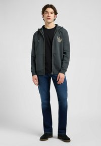 Felpa zip nera con cappuccio e logo frontale, abbinata a una t-shirt nera e jeans blu. Il modello sta in piedi con le mani sui fianchi, indossando sneakers.
