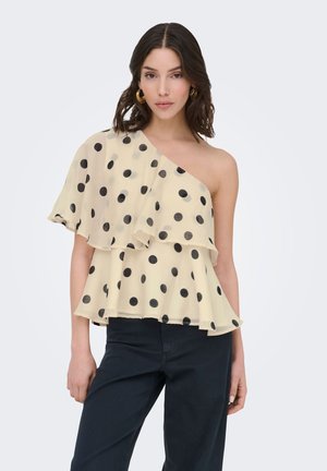 Kvinde iført en beige bluse med ét skulderstykke med sorte polkaprikker og flæselag, kombineret med mørke bukser, stående foran en ensfarvet baggrund.