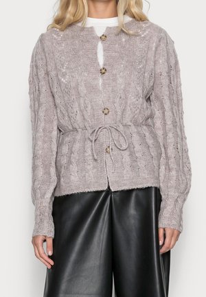 Kvinde iført en lavendel farvet kabelstrikket cardigan med brune knapper, bundet i taljen, over en hvid skjorte og sorte læderbukser.