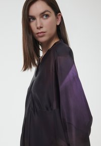 Zijdeachtige donkerpaarse blouse met een V-hals en lange, brede mouwen. Heeft een vloeiende ontwerp en subtiele kleurvariaties.