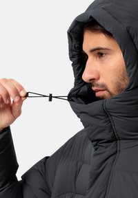 Veste matelassée noire avec capuche, tissu léger, texture matelassée, cordon ajustable, gros plan sur une main saisissant le toggle.