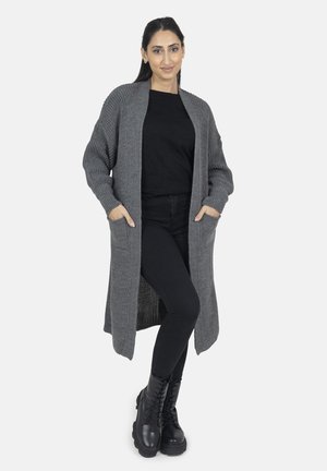 Graue lange Strickjacke aus strukturiertem Strickstoff, mit zwei vorderen Taschen und einem lässigen Schnitt, getragen über einem schwarzen Oberteil und einer Hose. Schwarze Schnürstiefel.