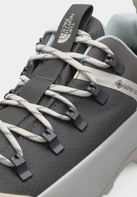 Κοντινό πλάνο γκρι και λευκού αθλητικού παπουτσιού North Face με κορδόνια με σχέδιο, ετικέτα γλώσσας με λογότυπο και ετικέτα αδιάβροχου Gore-Tex στο πλάι.