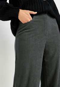 Pantalons en velours côtelé vert foncé avec une finition texturée, dotés de poches latérales et d'une coupe décontractée. Un pull en tricot noir les couvre partiellement.