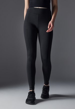 Leggings neri realizzati in tessuto elasticizzato con vita alta, caratterizzati da cuciture piatte e un design elegante. Indossati con scarpe da ginnastica nere.