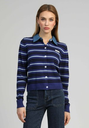 Femme portant un cardigan boutonné à rayures bleues avec un col en denim et un jean foncé, debout devant un fond uni gris clair.