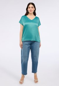 Tyrkysový saténový top s výstřihem do V, krátkými rukávy a volným střihem, doplněný o světlé modré džíny a bílé sandály na podpatku.