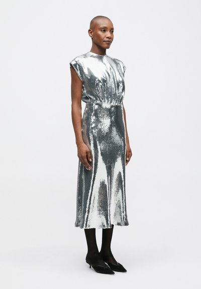 Sieviete, tērpusies sudraba spīguļotā midi kleitā ar plecu spilveniem, melnās zeķbiksēs un melnās smailējošās zemas papēža kurpēs, stāv uz balta fona.