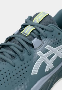 Σκούρο πετρόλ παπούτσι τρεξίματος Asics με γκρι κορδόνια, διάτρητη πλευρά, λευκό λογότυπο και γλωσσάκι με έντονες κίτρινες λεπτομέρειες.